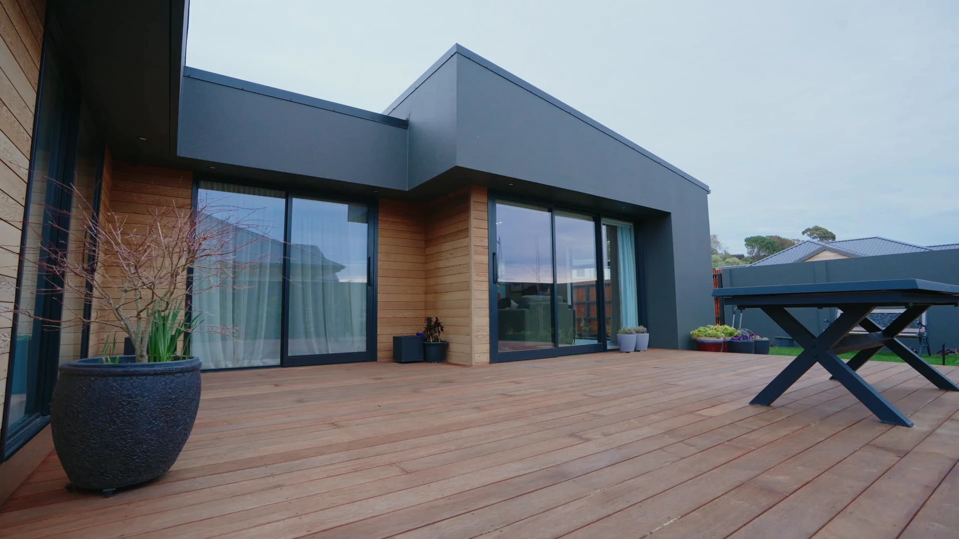 Charcoal render and cedar around a wide timber deck, sliding doors running the length of the living wing